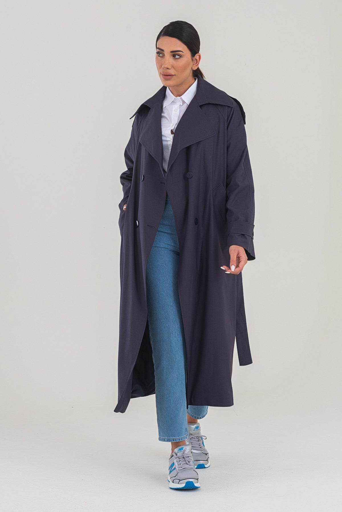 Woman wearing a long dark coat over a white shirt and blue jeans on a white background
