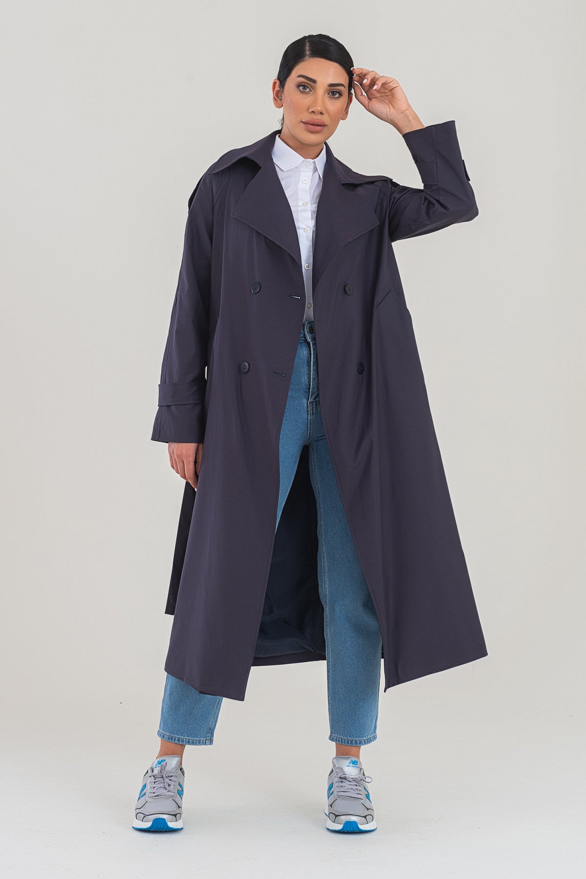 Person wearing a long dark coat over a white shirt and blue jeans on a plain background