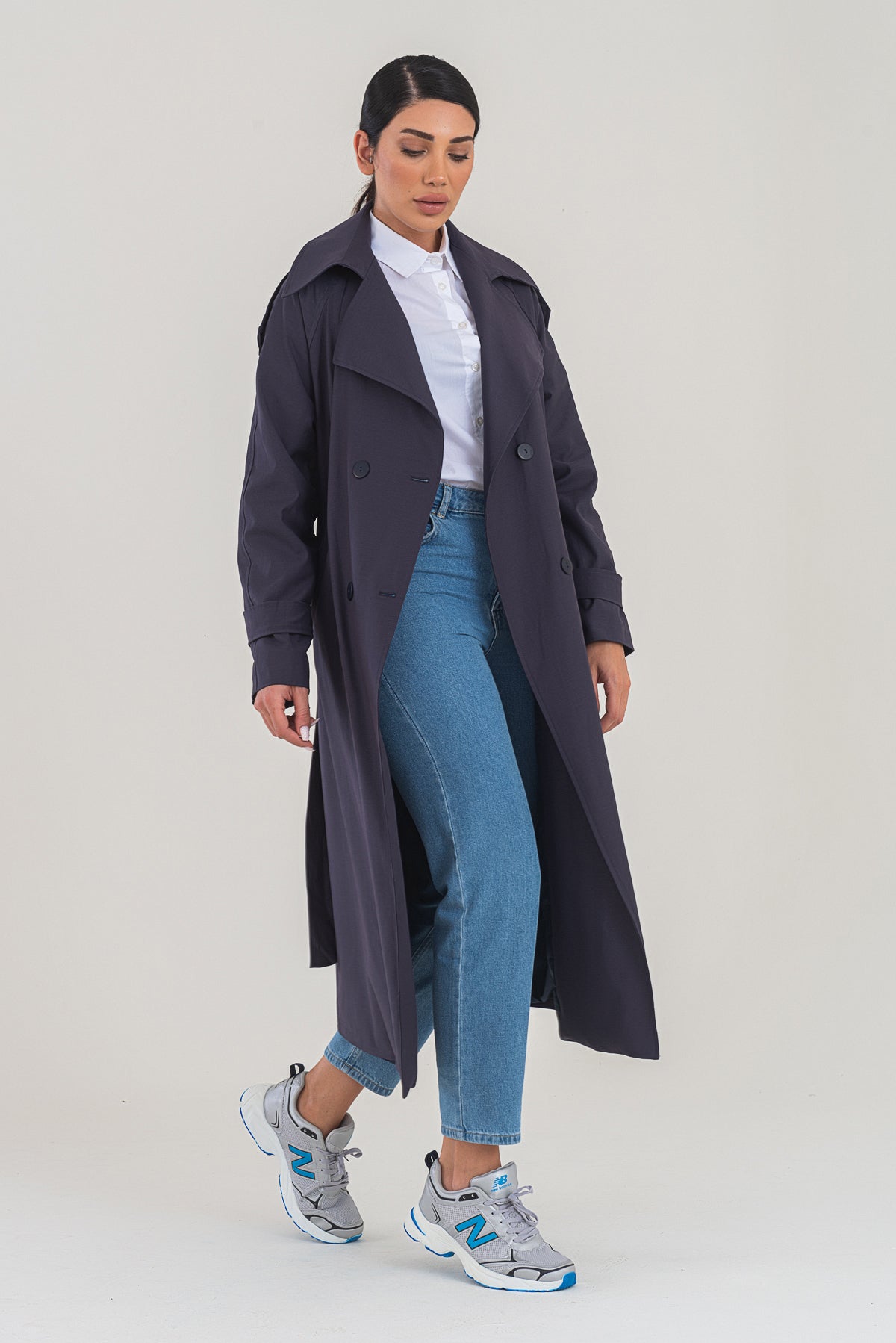 Woman wearing a long dark coat, white shirt, blue jeans, and gray sneakers on a light background