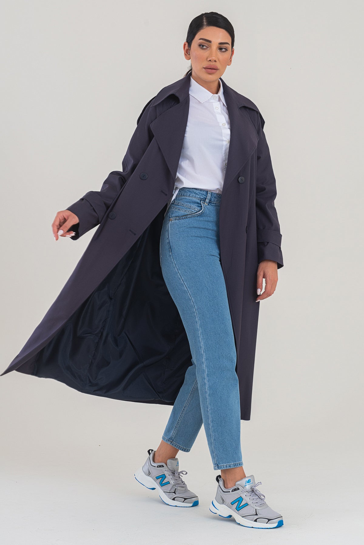 Woman wearing a long dark coat, white shirt, blue jeans, and gray sneakers on a light background