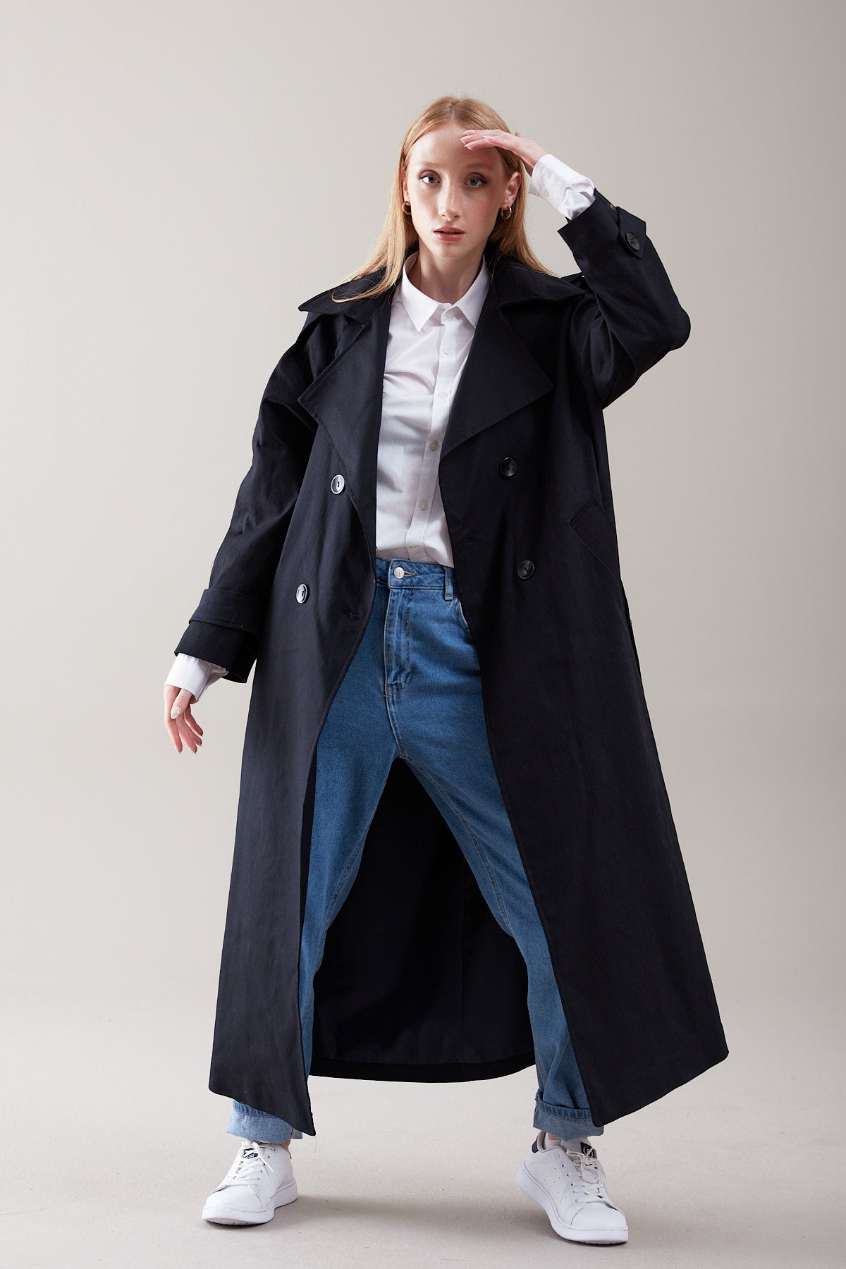 Person wearing a long black coat, white shirt, blue jeans, and white sneakers on a plain background