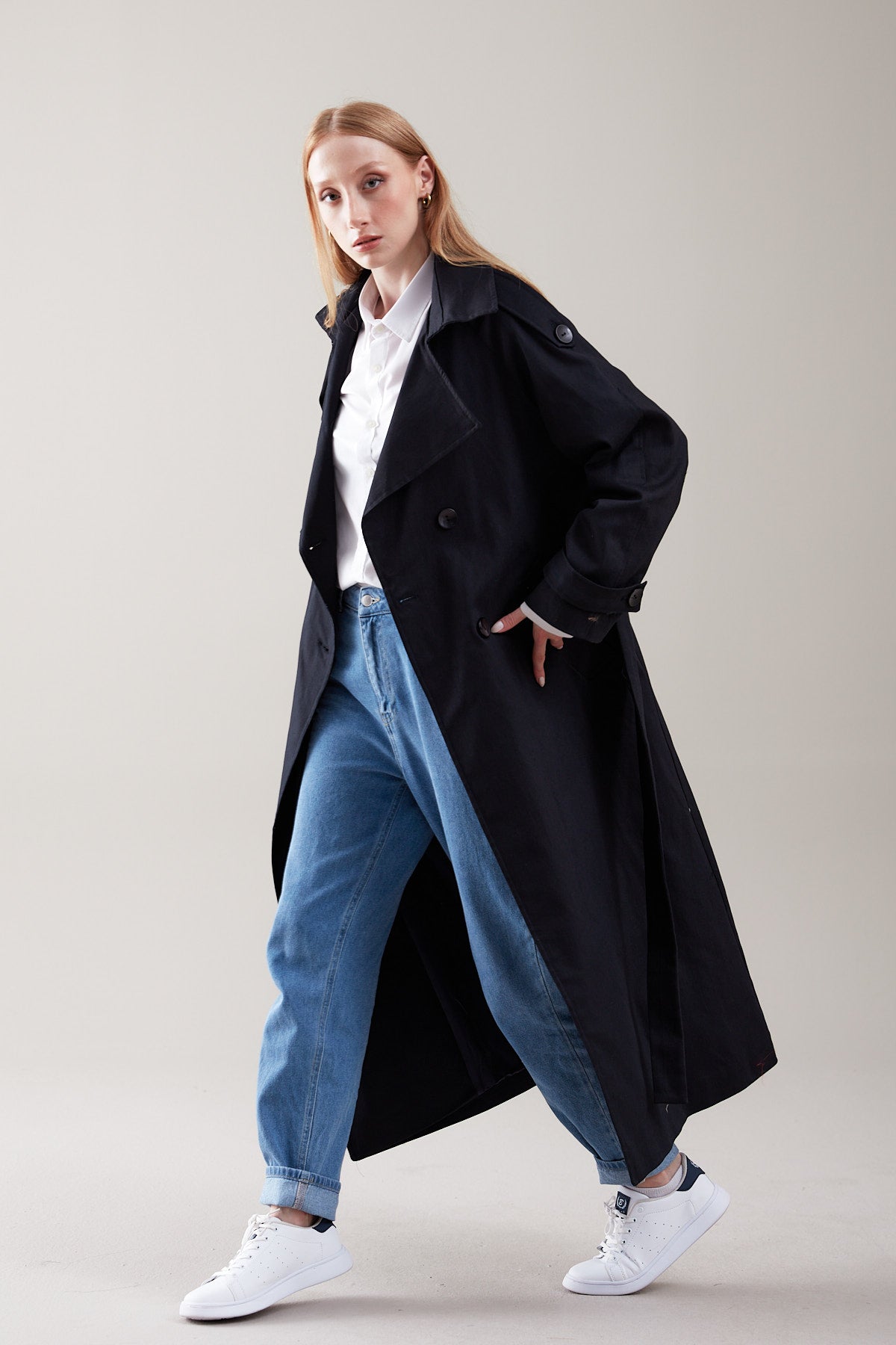 Person wearing a black coat, white shirt, blue jeans, and white sneakers on a plain background